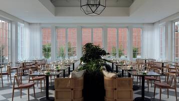 A spacious dining area with high ceilings, large windows, and a central plant. Tables are set with chairs, and the floor has a checkered pattern.