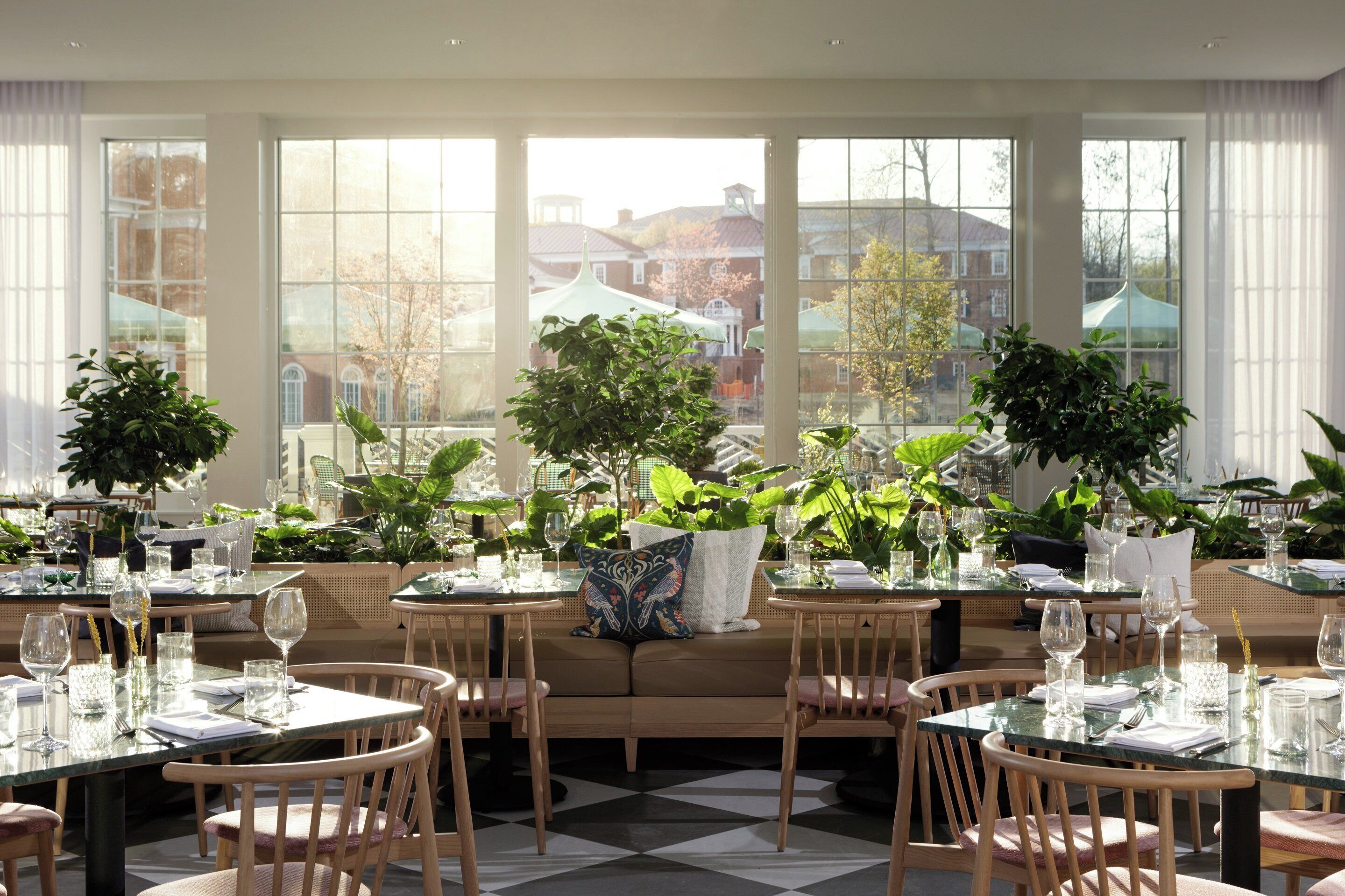 Un restaurant avec des tables dressées pour le service, de grandes fenêtres et des plantes d’intérieur.
