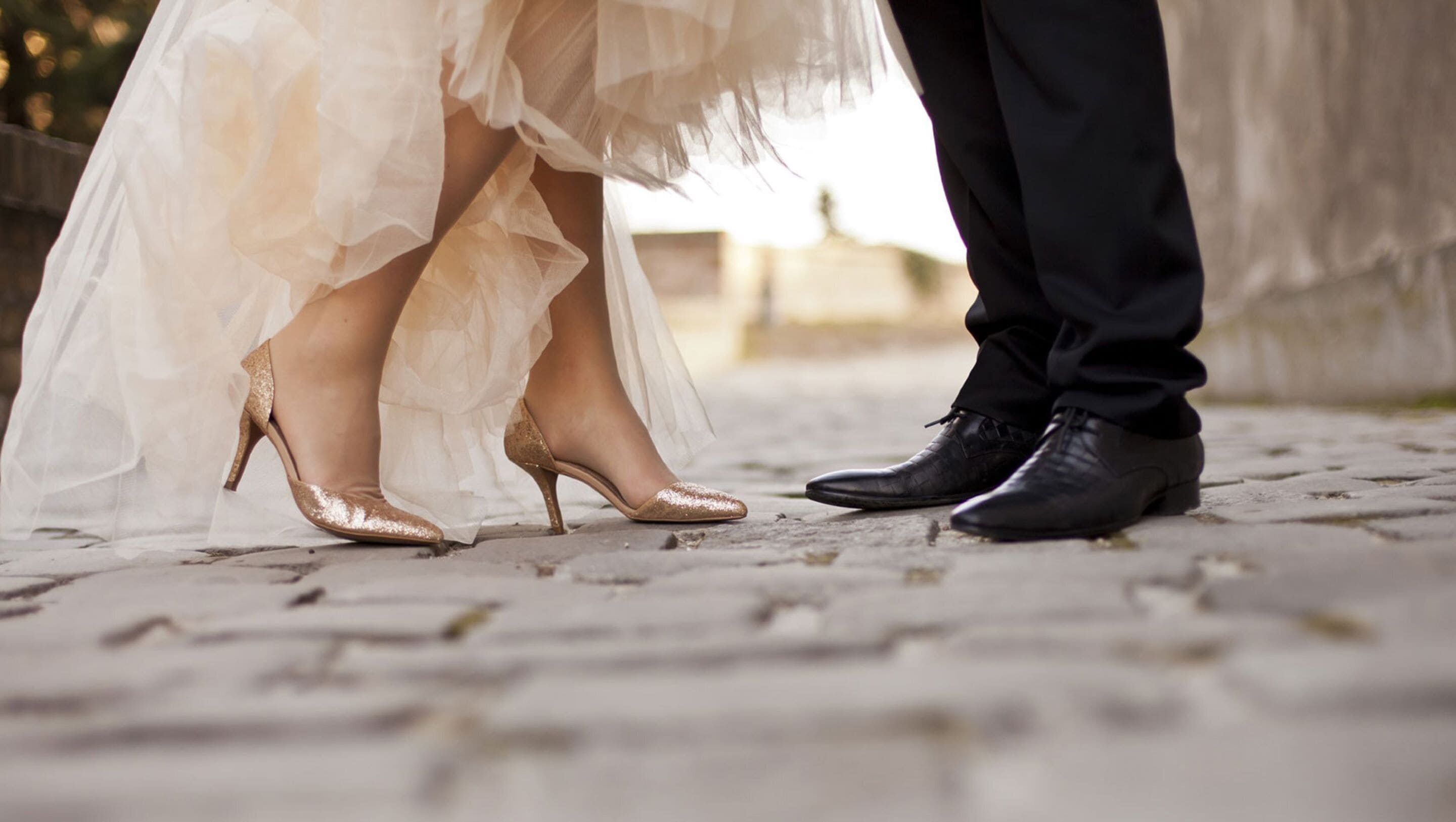 A close-up of a bride's feet in a white dress and high heels, and a groom's feet in black dress shoes on a cobblestone path.
