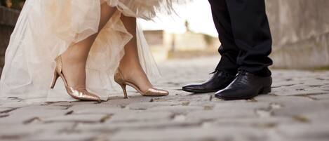 A close-up of a bride's feet in a white dress and high heels, and a groom's feet in black dress shoes on a cobblestone path.
