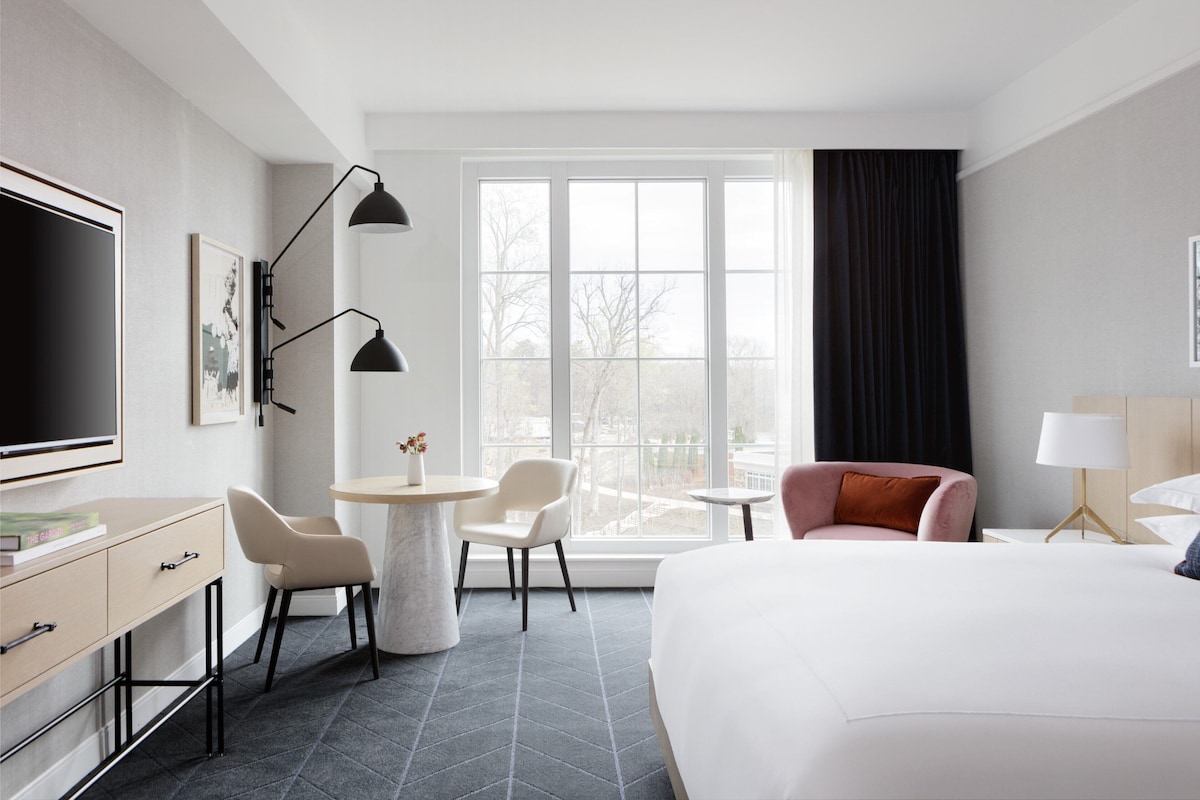 A hotel room with a bed, a desk, two chairs, a TV, and a large window with a view.