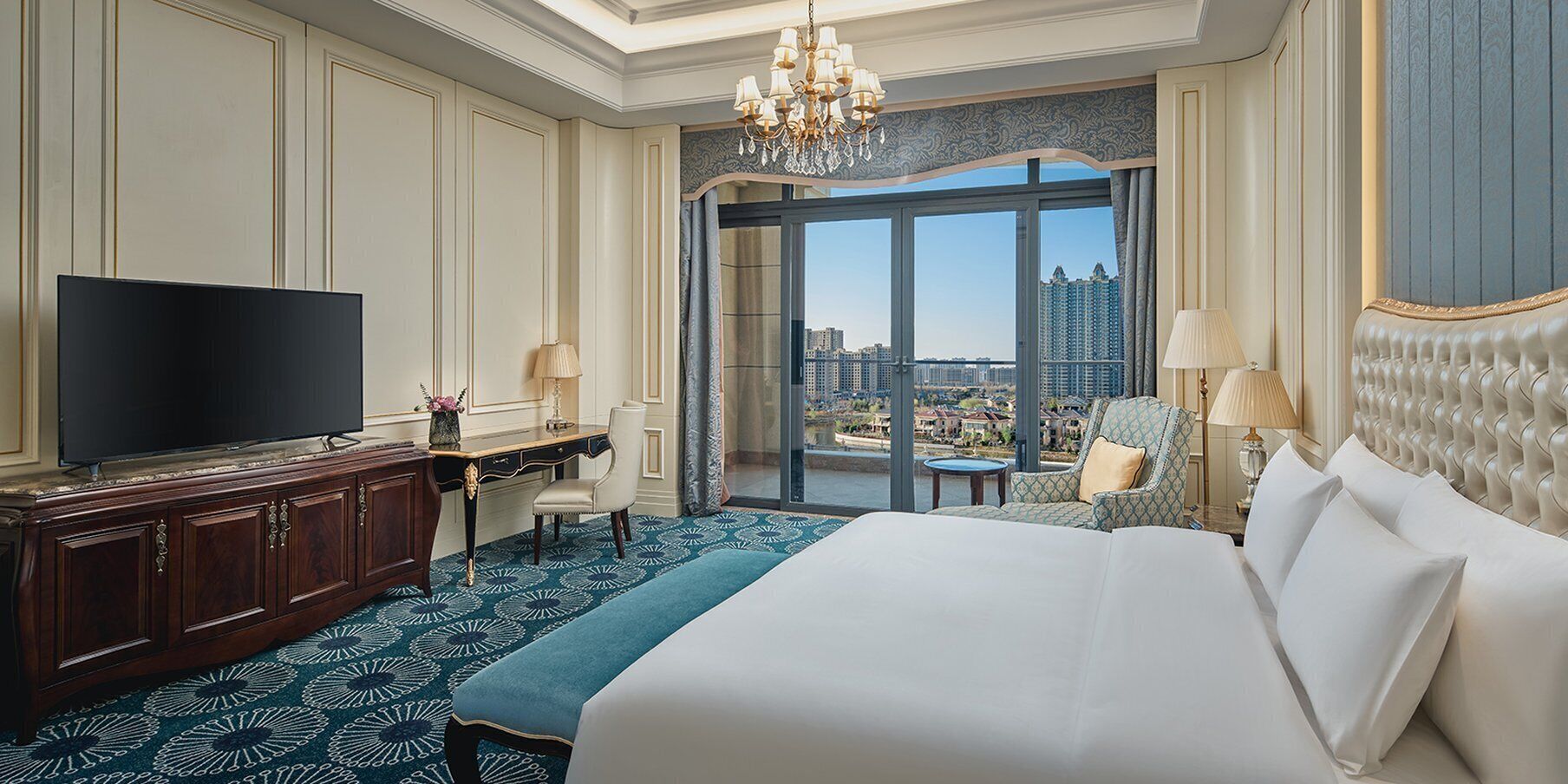 Ein Hotelzimmer mit einem großen Bett, einem Fernseher, einem Schreibtisch mit Stuhl und Blick auf eine Stadtlandschaft durch ein großes Fenster.