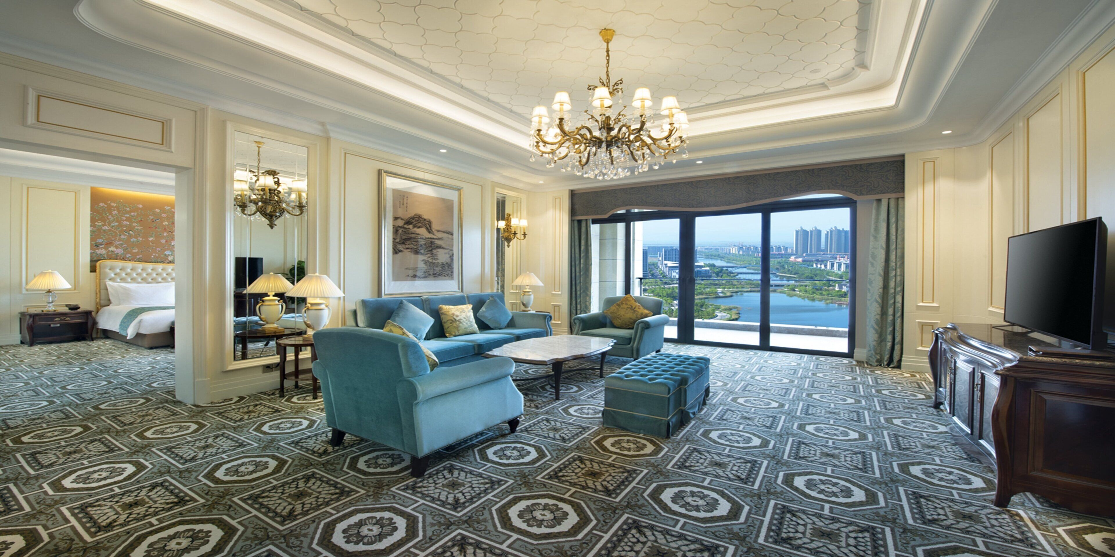 A hotel room with a large window, a chandelier, a television, and a patterned carpet.