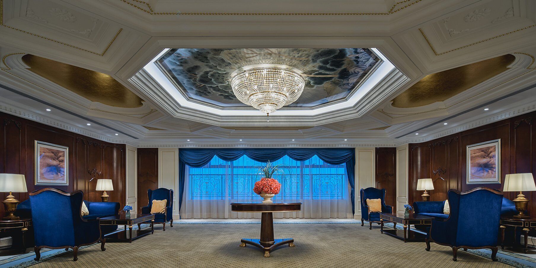 Un lobby lujoso con una imponente araña, una mesa central con un florero y sillones tapizados en azul.