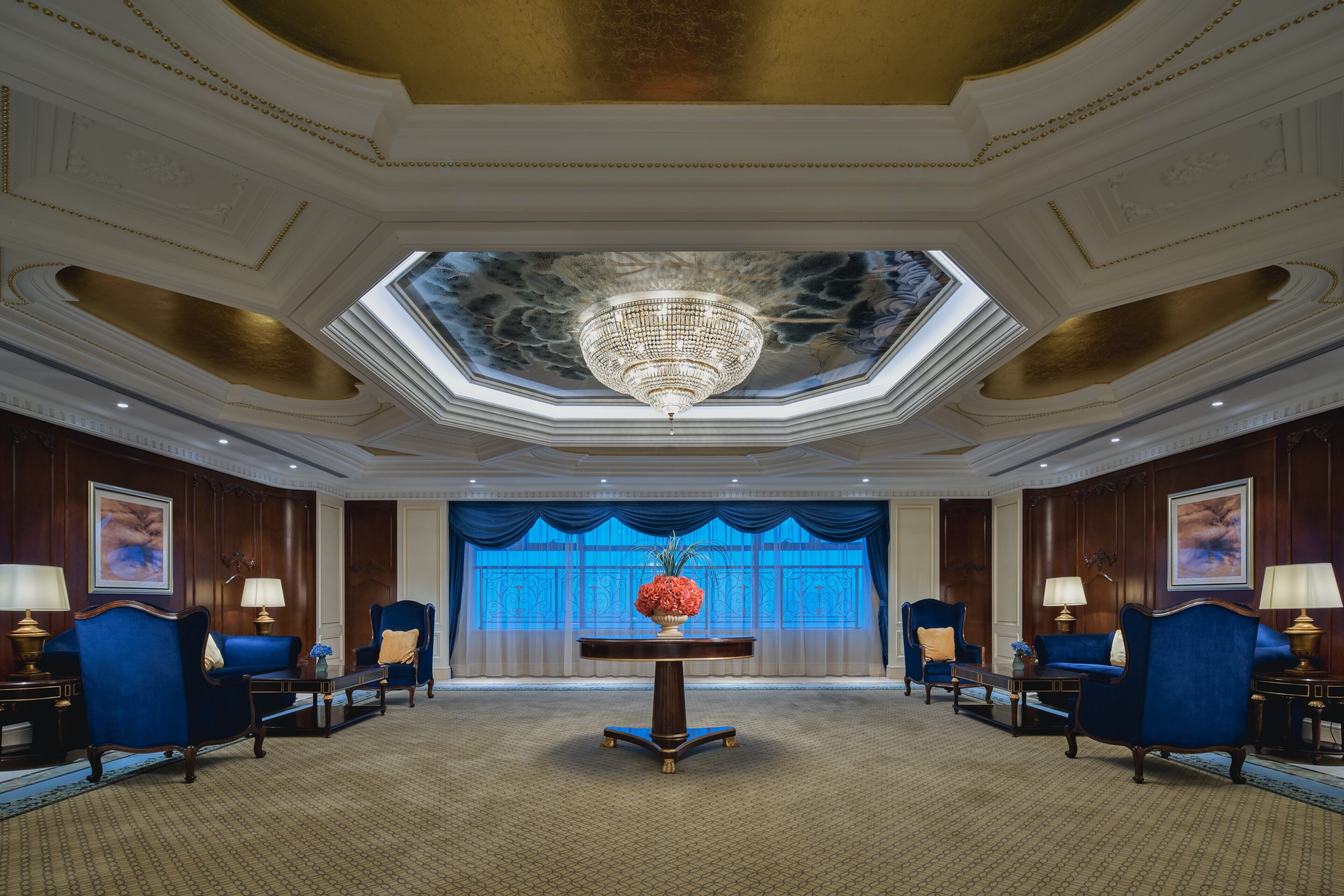 A grand lobby with a chandelier, blue upholstered chairs, a central table with a vase, and a large window with blue curtains.