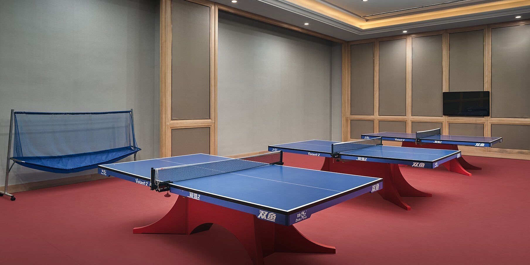 Quatre tables de ping-pong dans une pièce dotée d’un tapis rouge, d’une chaise bleue avec un filet et d’un téléviseur fixé au mur.