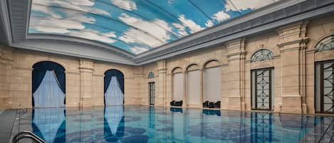 A large indoor swimming pool with a ceiling mural of clouds, arched doorways, and classical architecture.