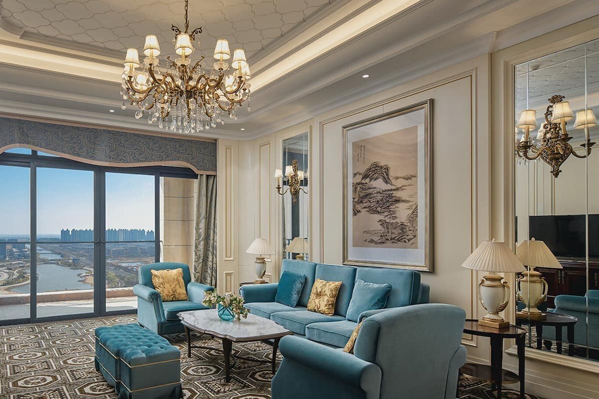 A spacious living room with a large TV, comfortable seating, a chandelier, and a view of a cityscape through large windows.
