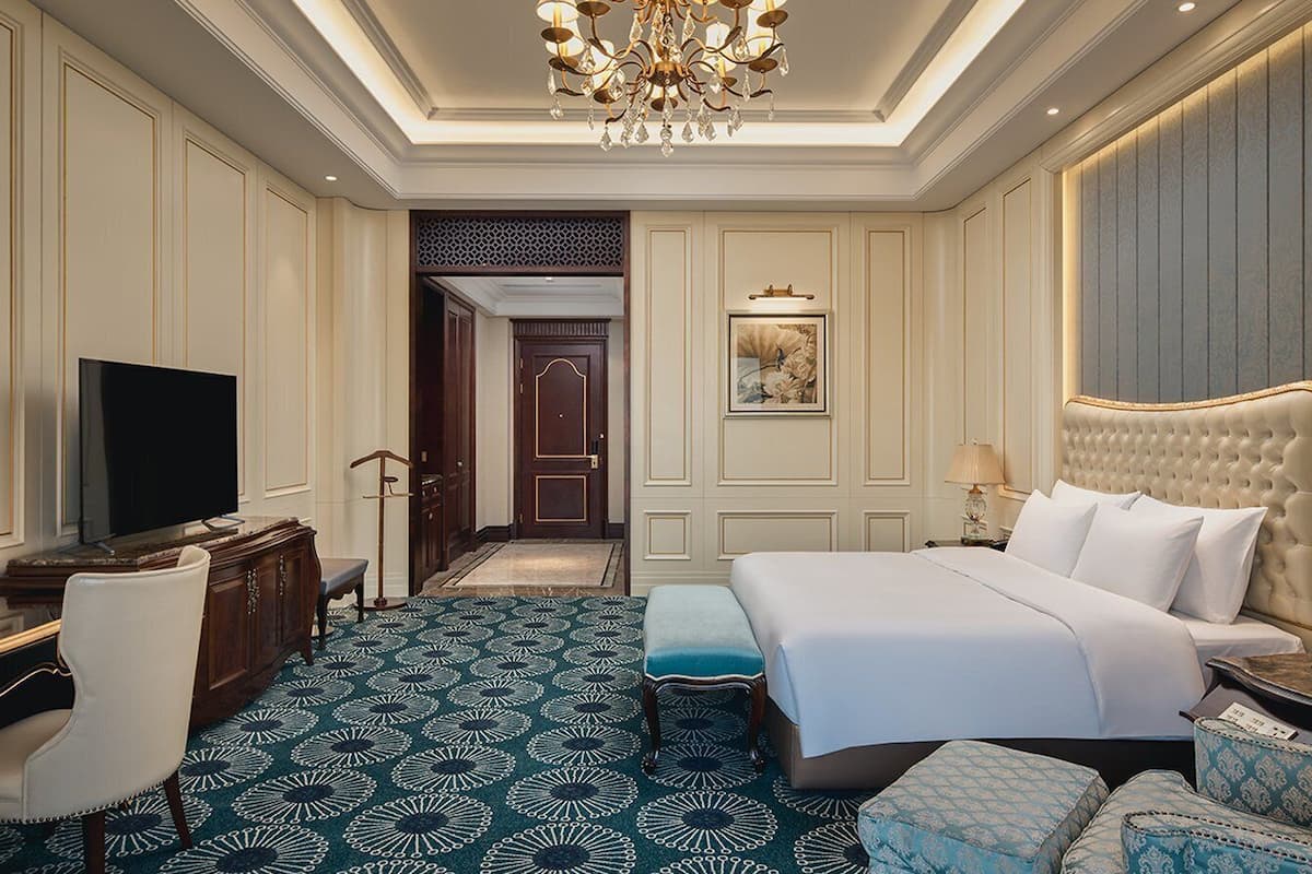 A hotel room with a large bed, a desk with a TV, a chair, and a patterned carpet.