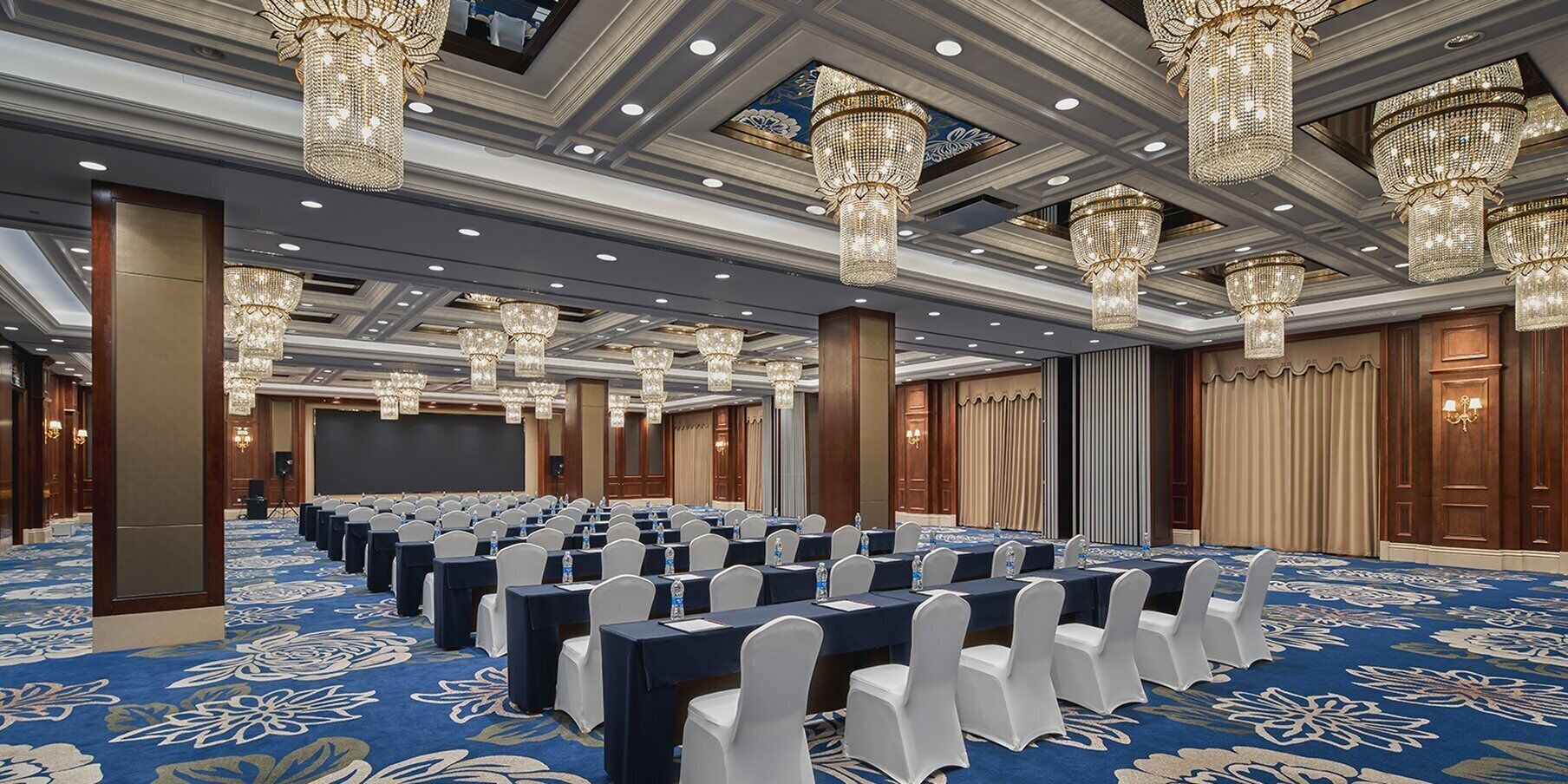 Een grote congreszaal met rijen tafels en stoelen, gedetailleerde kroonluchters en een blauw tapijt met bloemmotieven.