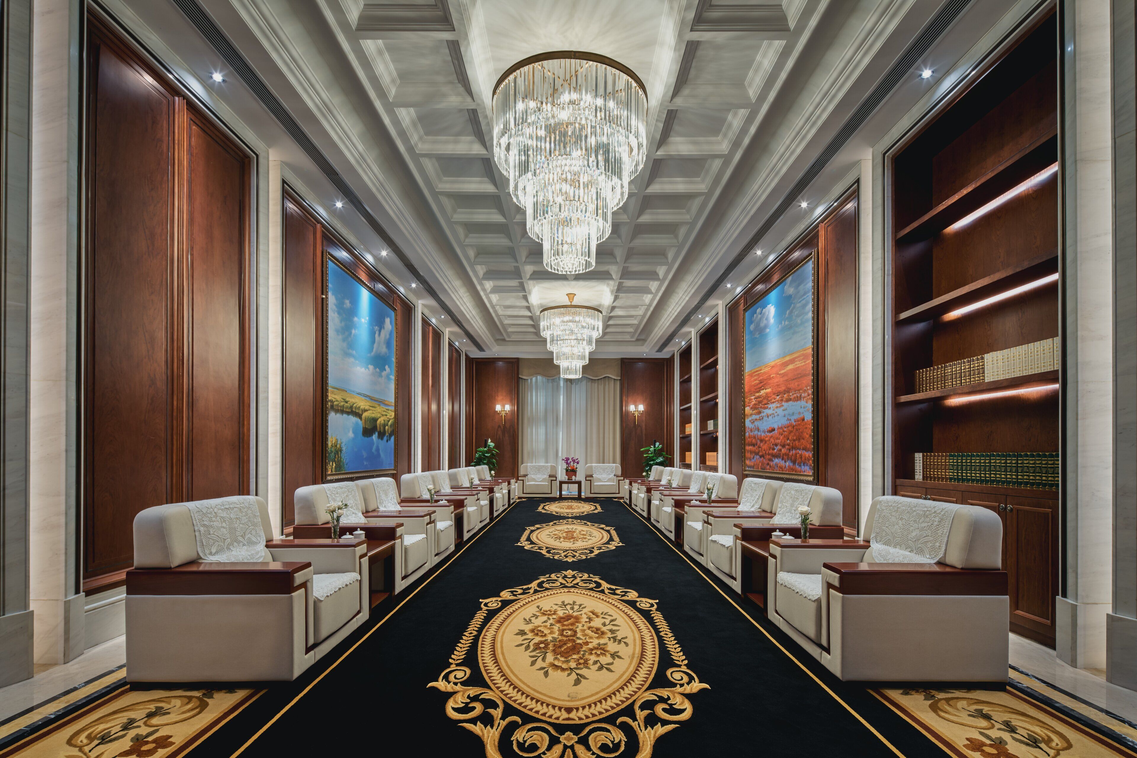 A hotel lobby with a central carpeted area, seating arrangements, chandeliers, and wall-mounted artwork.