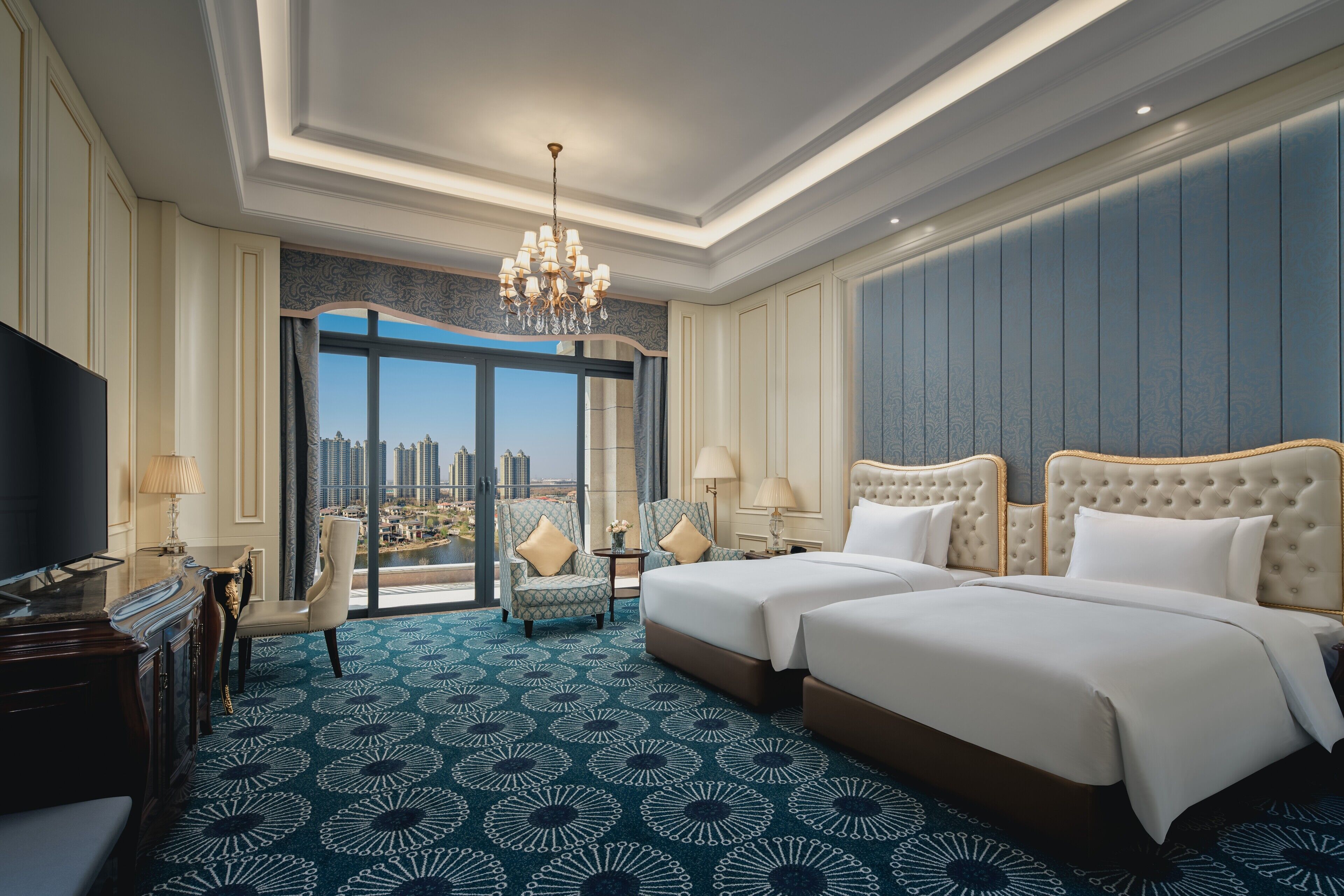 A hotel room with a large bed, two chairs, a desk, and a TV. The room has a view of a cityscape through a large window.