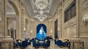 Een statige lobby met gedetailleerde kroonluchters, een vleugelpiano en blauwe fauteuils gerangschikt rond een centrale tafel.