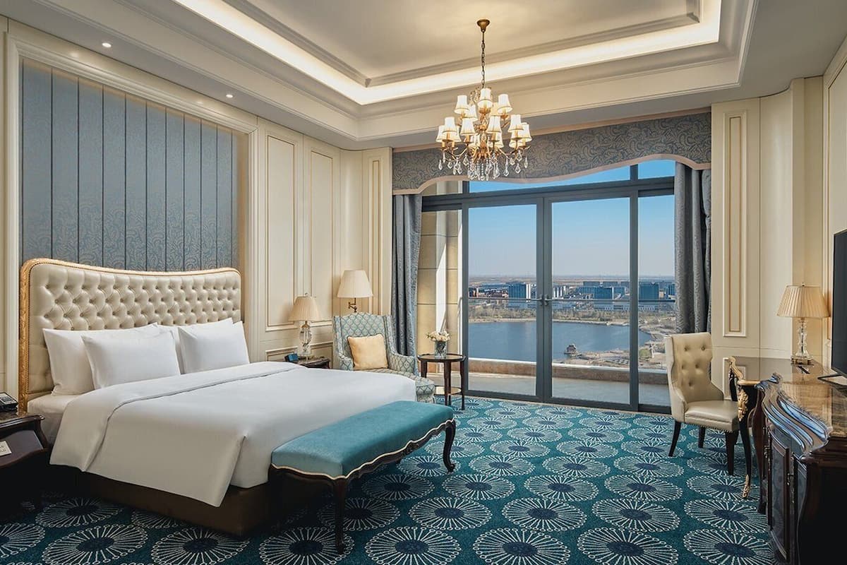 A hotel room with a large bed, a TV, a desk, and a view of a cityscape through large windows.