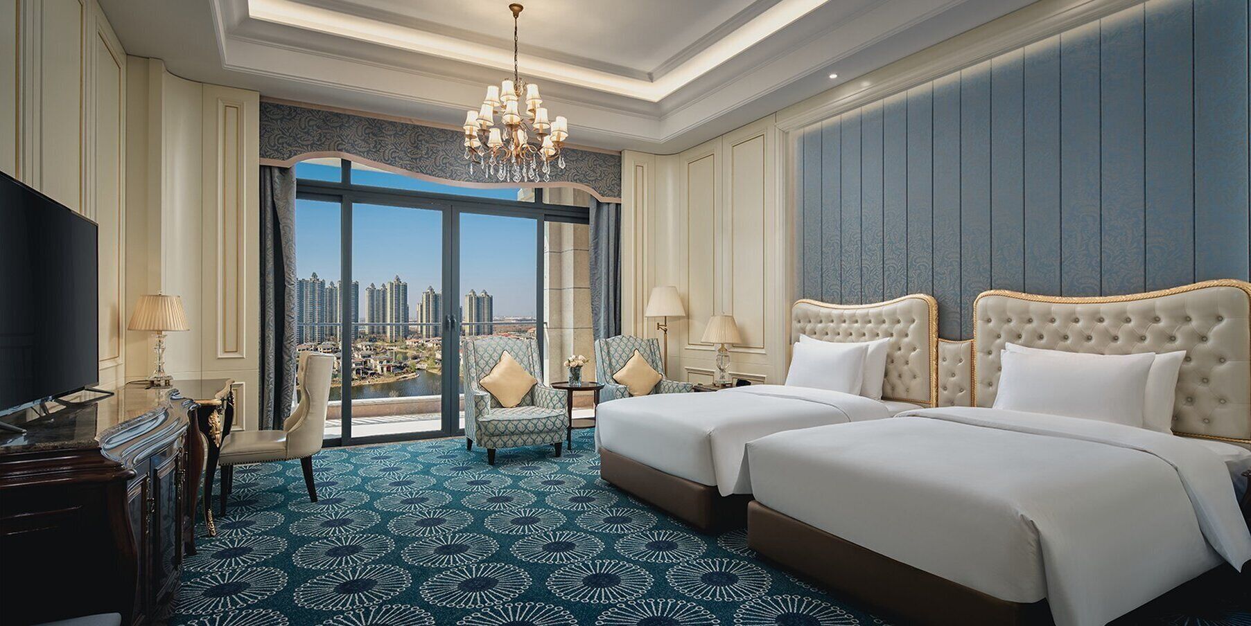 Ein Hotelzimmer mit einem großen Bett, zwei Nachttischen, einem Fernseher und Blick auf eine Stadtlandschaft durch große Fenster.