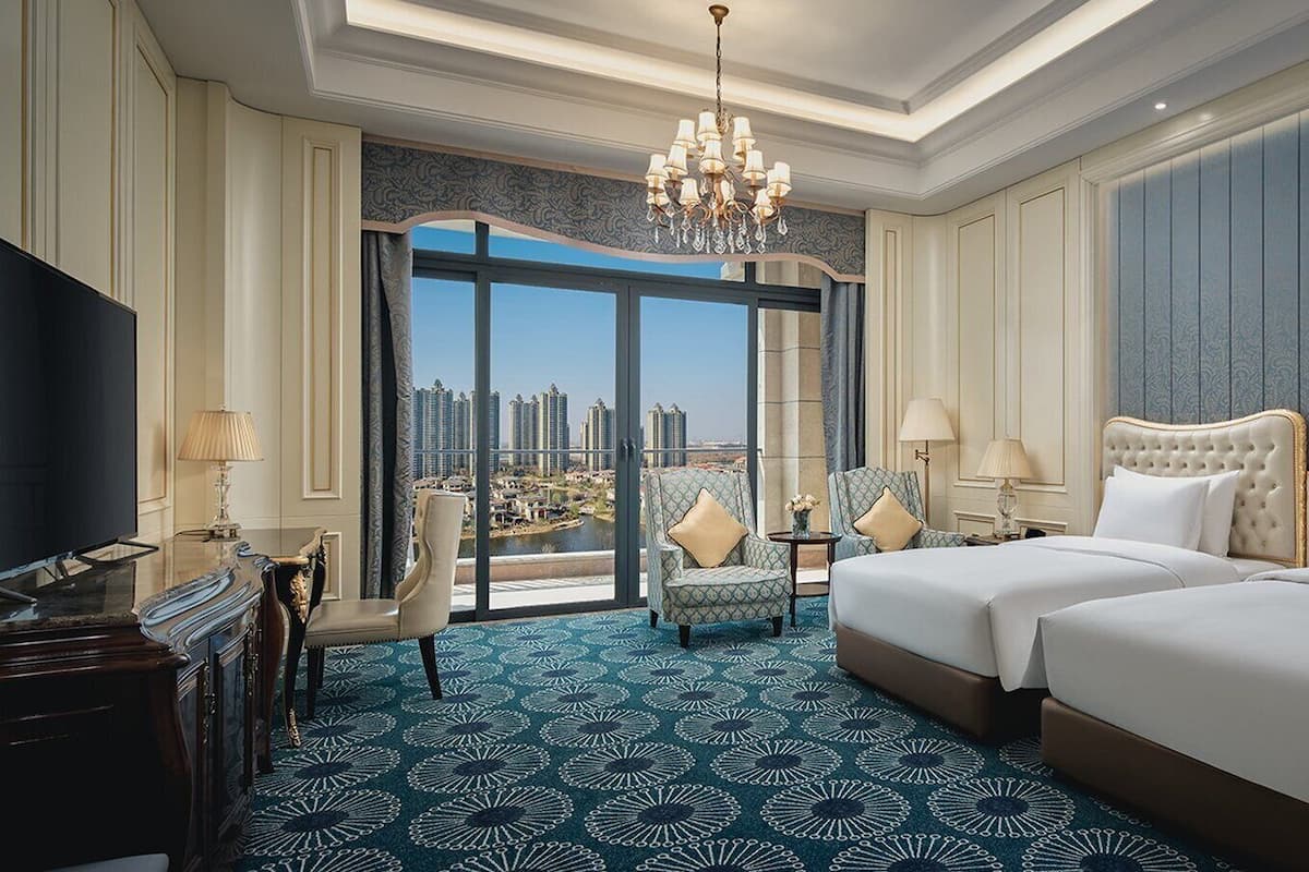 A hotel room with a large bed, two bedside tables, a TV, and a view of a cityscape through large windows.