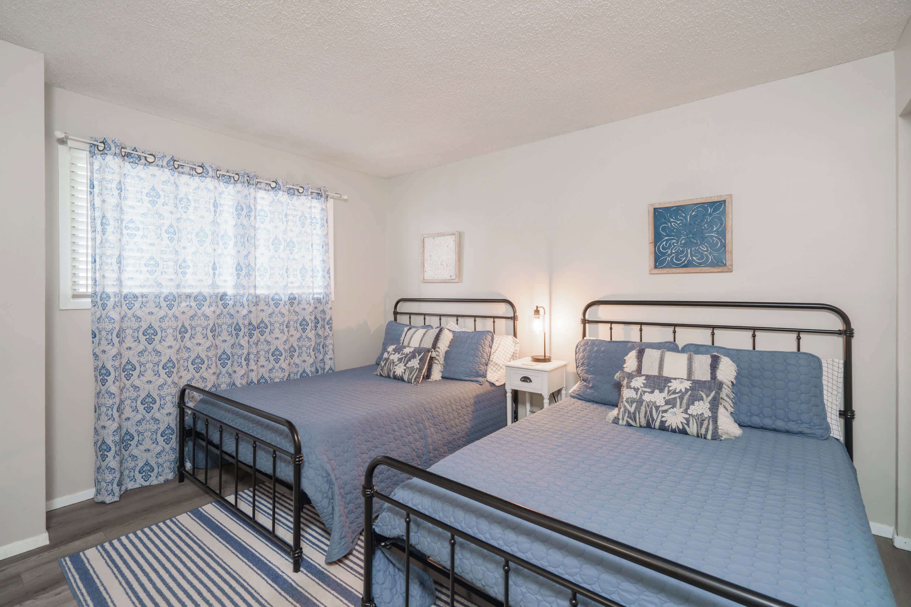 Un dormitorio con dos camas individuales, una mesita de noche, una ventana con cortinas estampadas y un cuadro colgado en la pared.