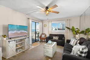 A living room with a television, a ceiling fan, a sofa, a coffee table, and a view of the ocean through a sliding door.