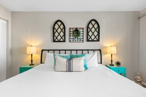Una cama bien tendida con edredón blanco y varios almohadones, flanqueada por dos mesitas de noche con lámparas, frente a una pared lisa con dos marcos decorativos que simulan ventanas.