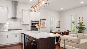 Una cocina moderna con armarios blancos, una isla central, electrodomésticos de acero inoxidable y una zona de comedor con mesa y sillas de madera.