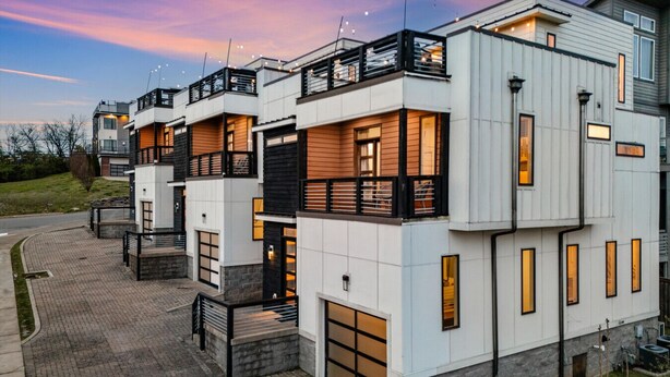 A modern multi-story residential building with a brick-paved driveway, large windows, and a balcony.