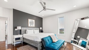 A modern bedroom with a bed, bedside tables, a ceiling fan, a large mirror, and a window with blinds.