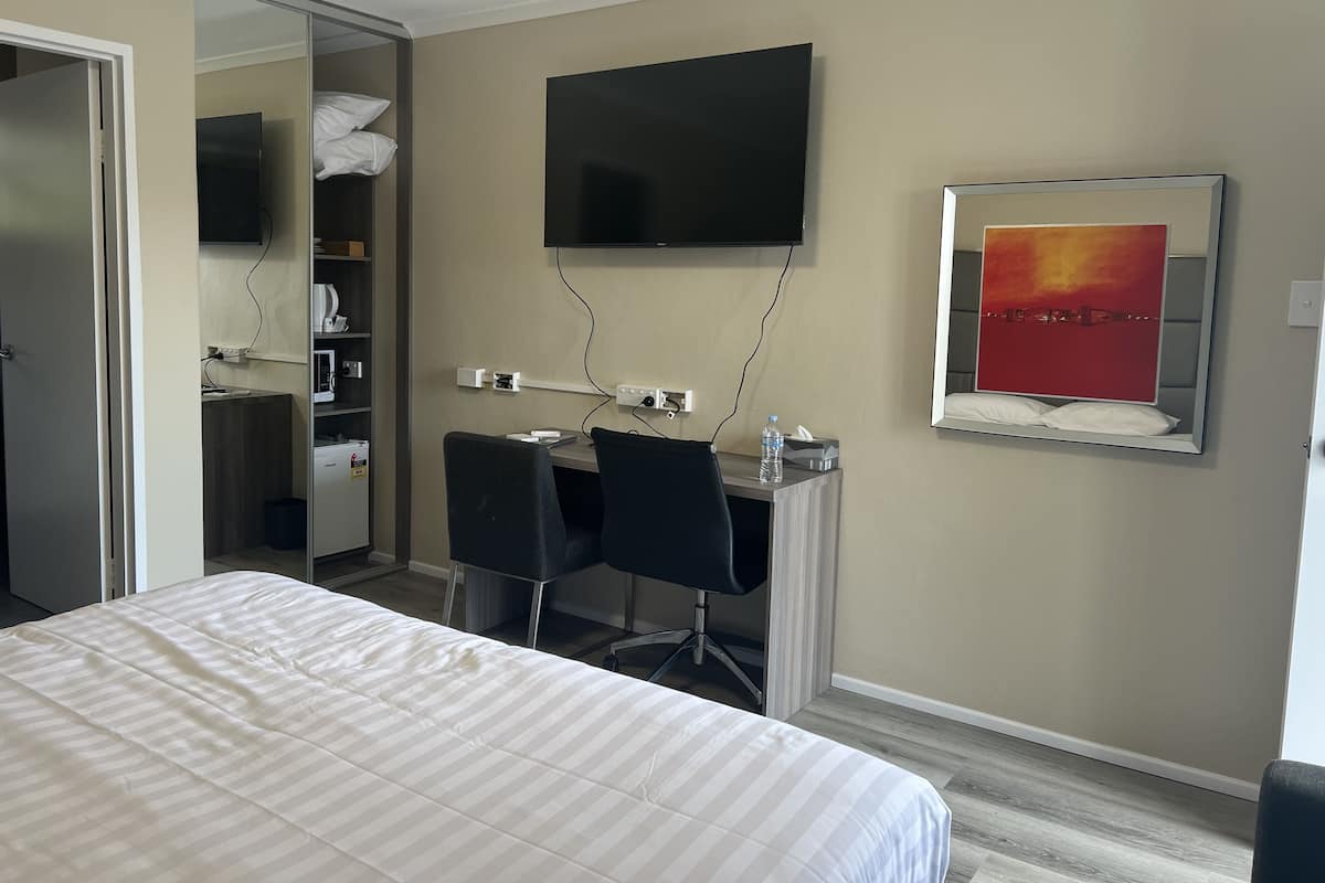 A hotel room with a bed, a desk with a chair, a television, and a mirror with a painting.
