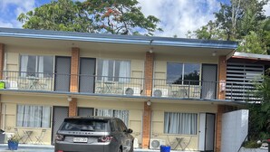 Un edificio de dos pisos con estacionamiento, un automóvil y un árbol con flores rojas.