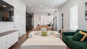 A modern living room with a sofa, a green couch, a kitchen in the background, and a dining area with a table and chairs.