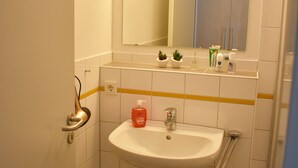 Ein kleines Badezimmer mit einem weißen Waschbecken, einem Spiegel, einem kleinen Hocker und einem blauen Mülleimer.