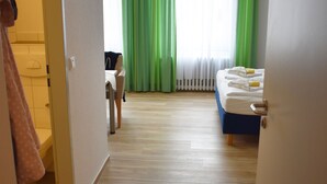 Ein Hotelzimmer mit Holzboden, einem Bett mit weißer Bettwäsche, einem Schreibtisch mit Stuhl und einem Fenster mit grünen Vorhängen.