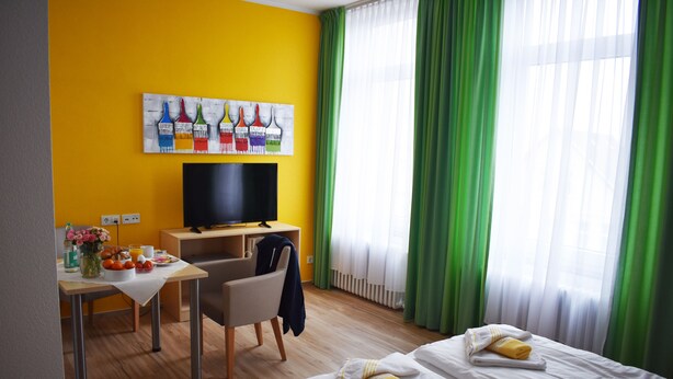 Ein Hotelzimmer mit gelber Wand, einem Gemälde von Pinseln, einem Schreibtisch mit Stuhl, einem Bett mit Handtüchern und einem Fernseher.