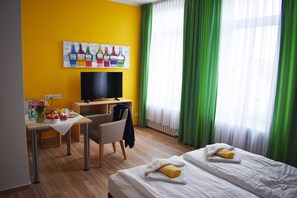 A hotel room with a yellow wall, a painting of paintbrushes, a desk with a chair, a bed with towels, and a TV.