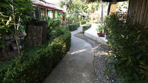 A pathway leading through a lush garden area with a thatched-roof structure and potted plants. - Avocado (Koh Samui)