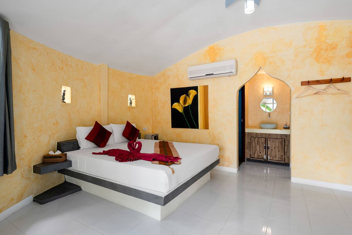 Een moderne hotelkamer met een groot bed, nachtkastjes, een airconditioning en een kleine badkamer met een wastafel en een spiegel.