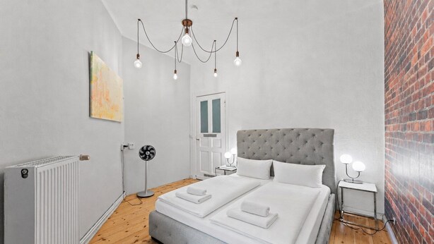 Une chambre avec un lit recouvert d’un tissu gris, un radiateur, un ventilateur sur pied et un mur en briques.