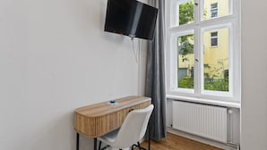 Une pièce avec un téléviseur fixé au mur, un bureau en bois avec une chaise, et une fenêtre donnant sur des bâtiments et de la verdure.
