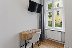 A room with a TV mounted on the wall, a wooden desk with a chair, and a window with a view of buildings and greenery.