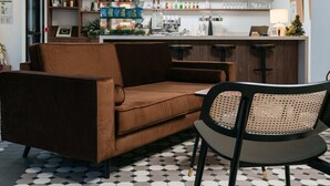 Et moderne barområde med en sofa og en lænestol i forgrunden, en bar med hylder fyldt med flasker og glas, samt en pendellampe i loftet.