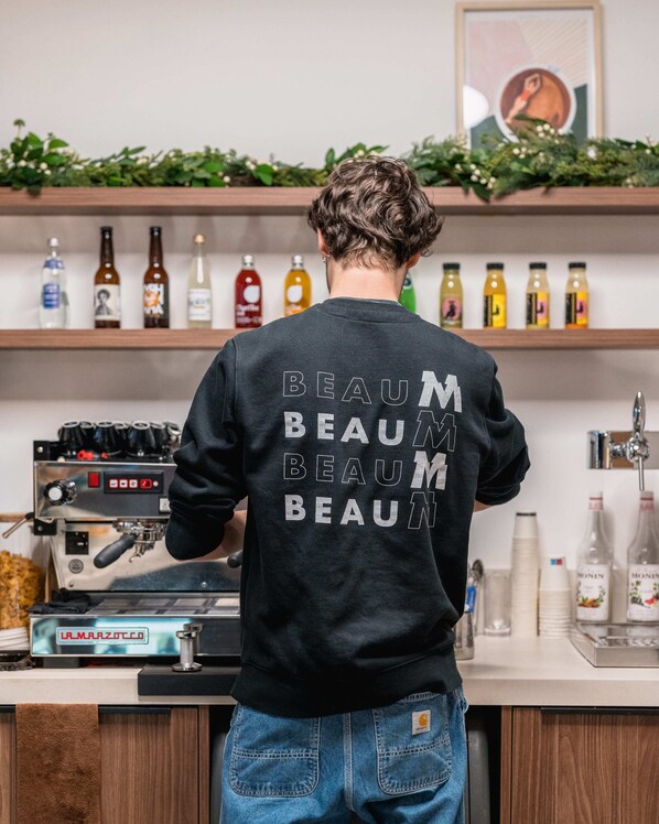 A person standing in front of a coffee machine, wearing a black sweatshirt with "BEAU BEAU BEAU" printed on the back.