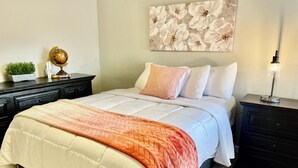 A neatly made bed with a white and peach quilt, a dark nightstand with a lamp, and a floral wall art piece above the bed. - AZ Peaceful Retreat in Scottsdale (Scottsdale)