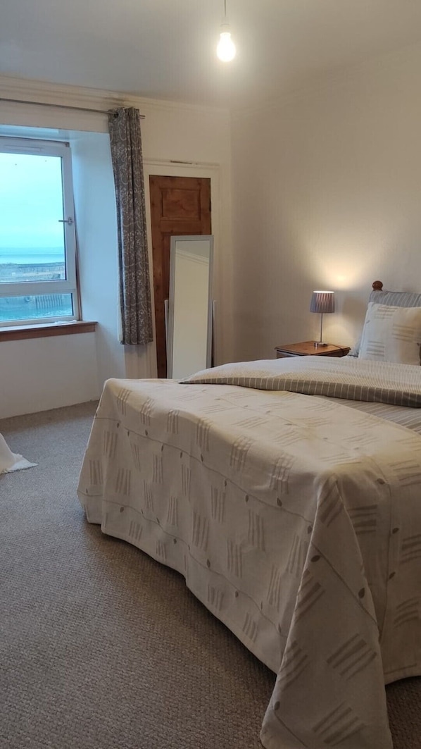 A bedroom with two beds, a window with a view, a wooden door, and a bedside lamp. - Kirkcaldy, Fife • Lovely 2 Bedroom Flat • Sea View (Kirkcaldy)