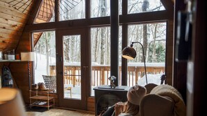 Een knus blokhutinterieur met een grote houten deur die uitkomt op een terras, een open haard en uitzicht op een besneeuwd bos.