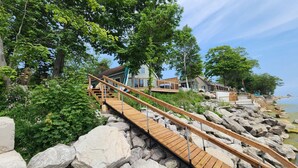 A wooden boardwalk leading to a house with a balcony, surrounded by rocks and greenery. - Lakefront Getaway | Hot Tub, Fire Pit & Sunsets (Selkirk)