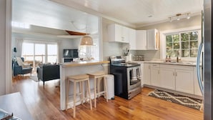 A modern kitchen with white cabinets, a stainless steel refrigerator, and a built-in oven. There are wooden stools and a large window providing natural light. - Lakefront Getaway | Hot Tub, Fire Pit & Sunsets (Selkirk)