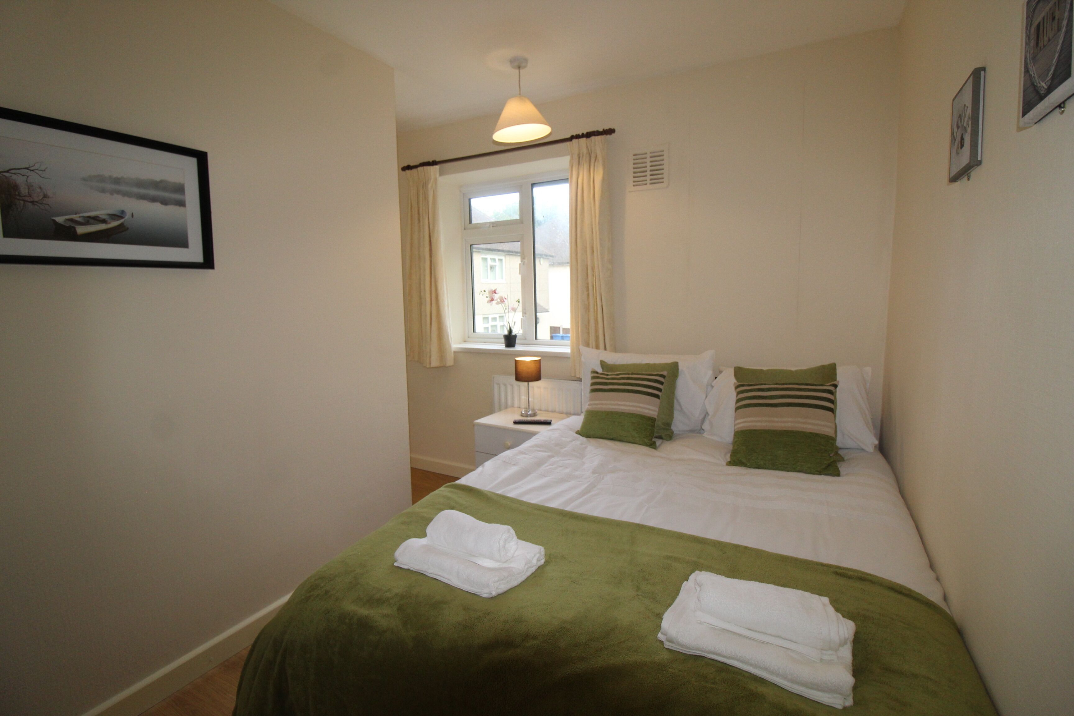 A neatly made bed with green and white bedding, a window with curtains, a bedside lamp, and a framed picture on the wall.