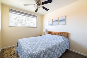 Une chambre à coucher comprenant un lit, un ventilateur de plafond, une fenêtre avec des stores et des œuvres d’art accrochées au mur.