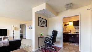 Una sala de estar moderna con un sofá, un escritorio de vidrio y una silla. Se ve una cocina a través de un vano abierto.