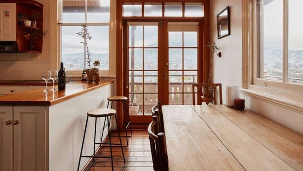 A kitchen with a wooden countertop, bar stools, a dining table, and a view through a large window.
