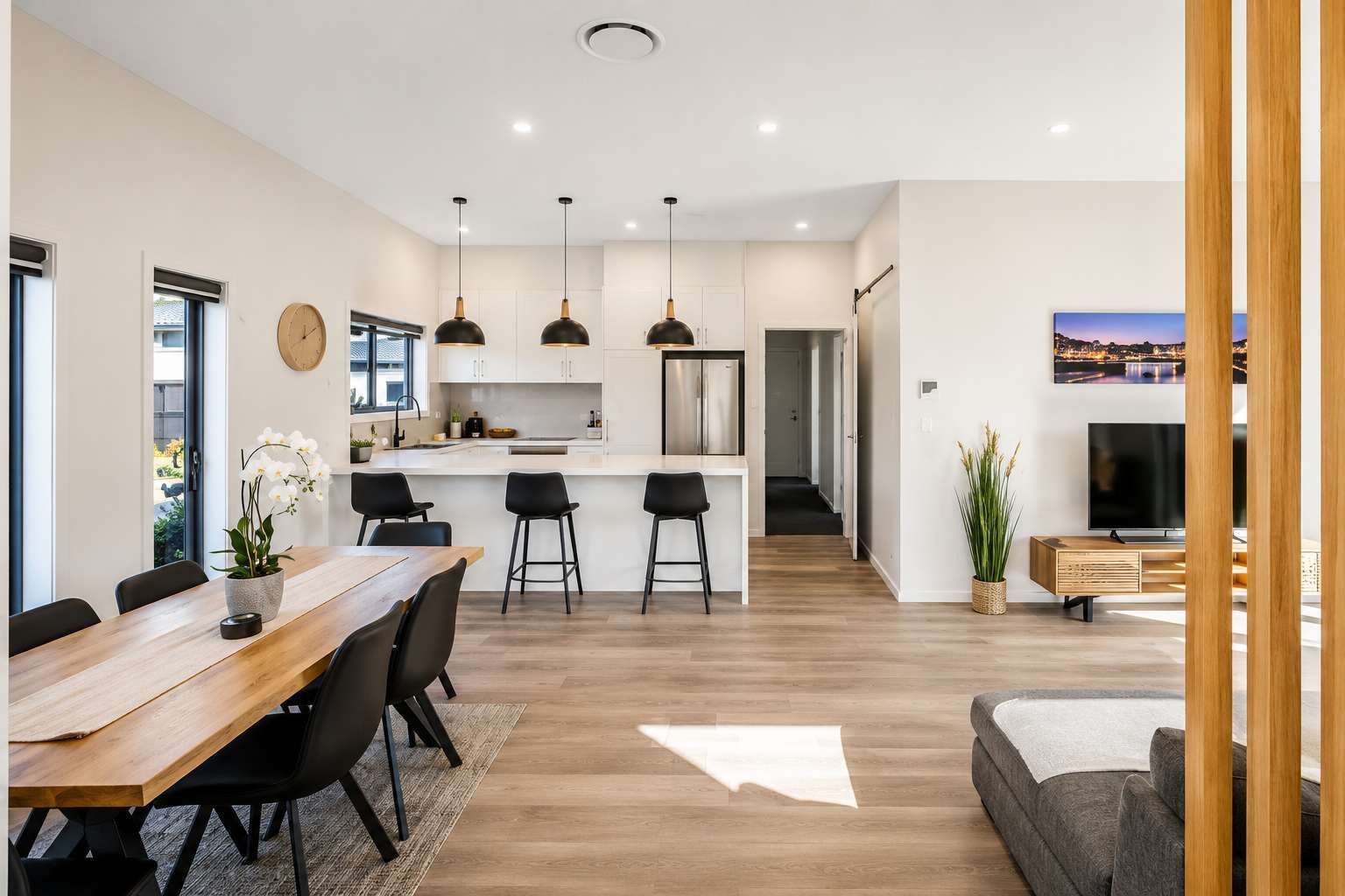 A modern kitchen with pendant lighting, a dining area with a wooden table and chairs, and a living room with a sofa and a TV.